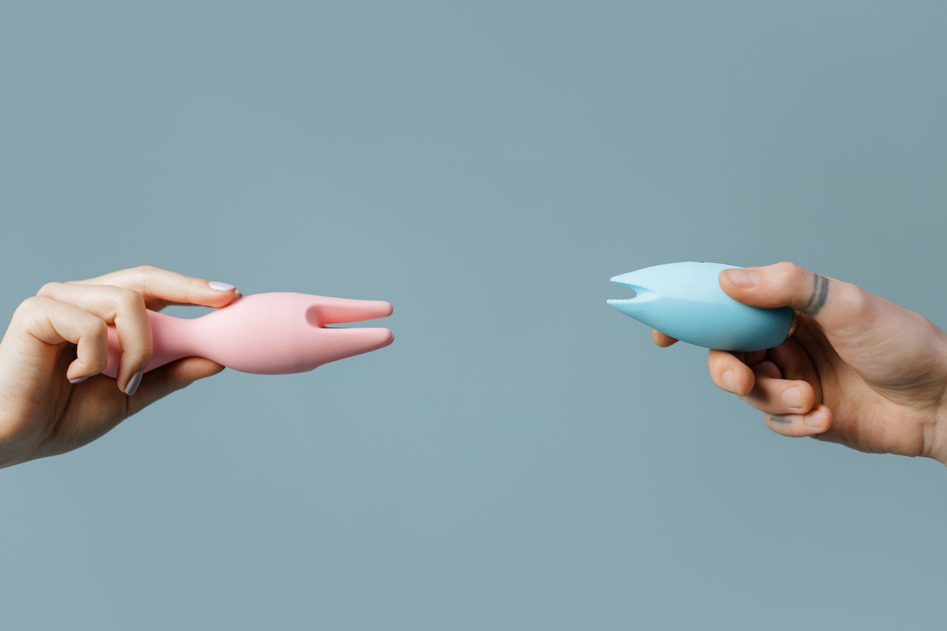 A hand holding a pink and blue silicone vibrator against a pastel background