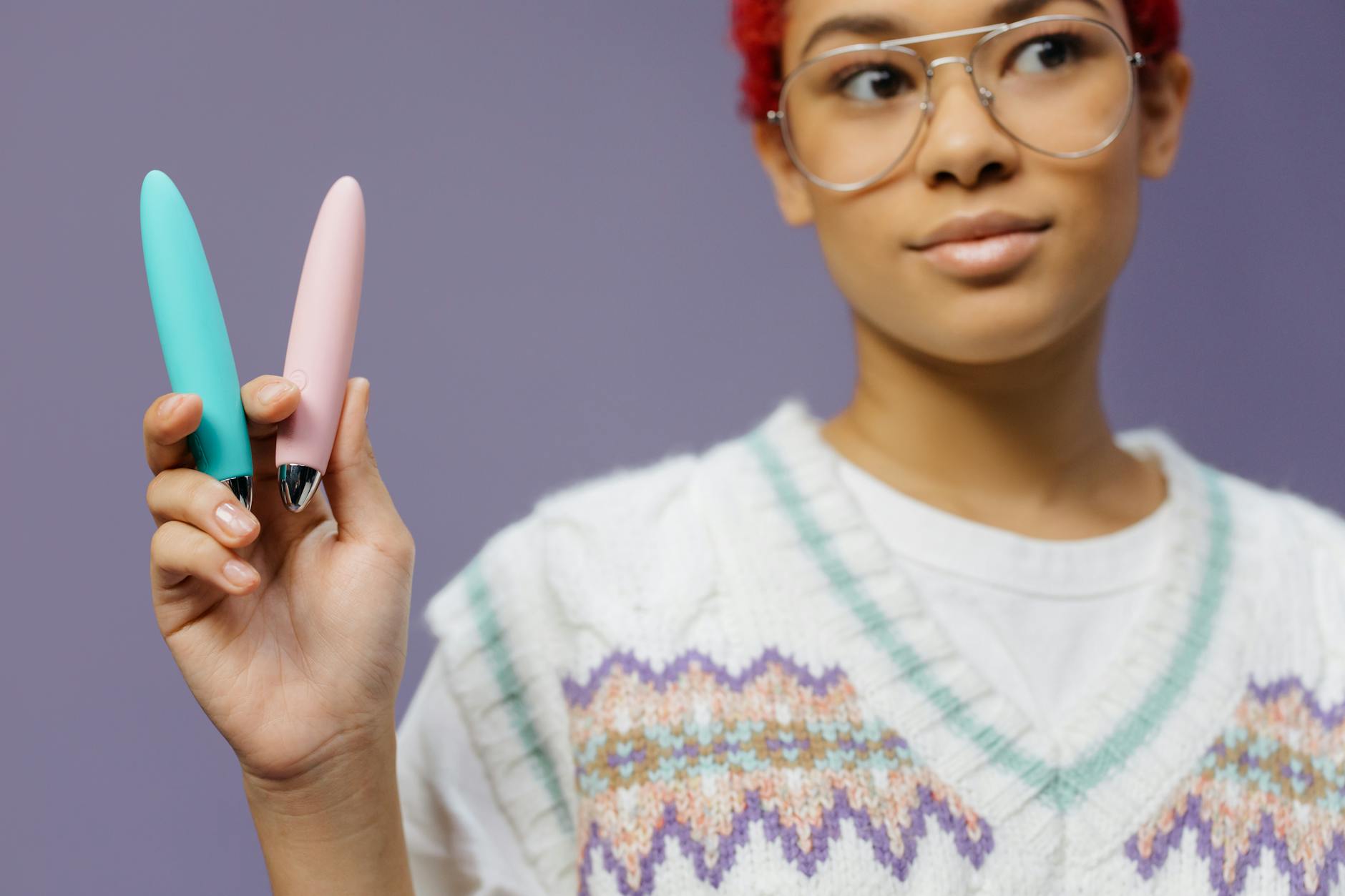 A young woman holding colorful vibrators against a purple background, representing personal pleasure without shame